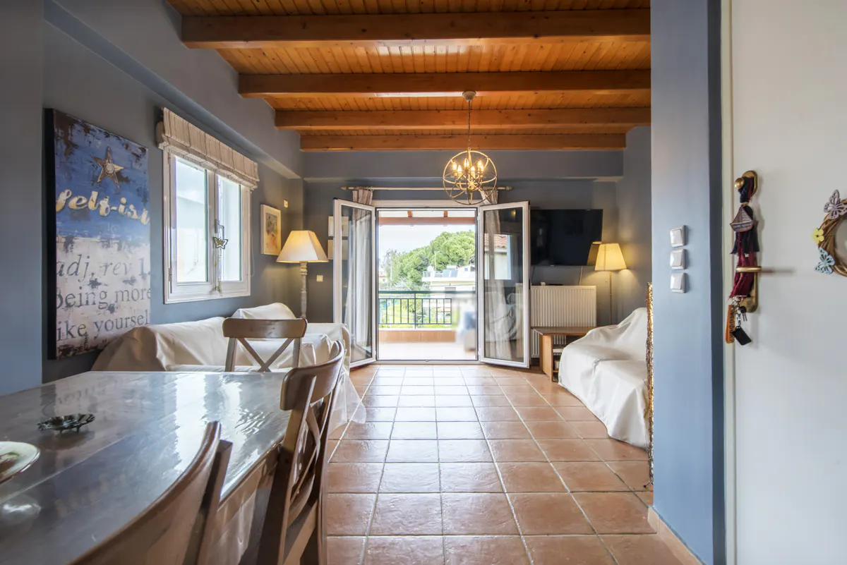 Living room with wood ceiling, tile floor, and open balcony doors. A dining table is on the left, and sofas are on either side of the room.