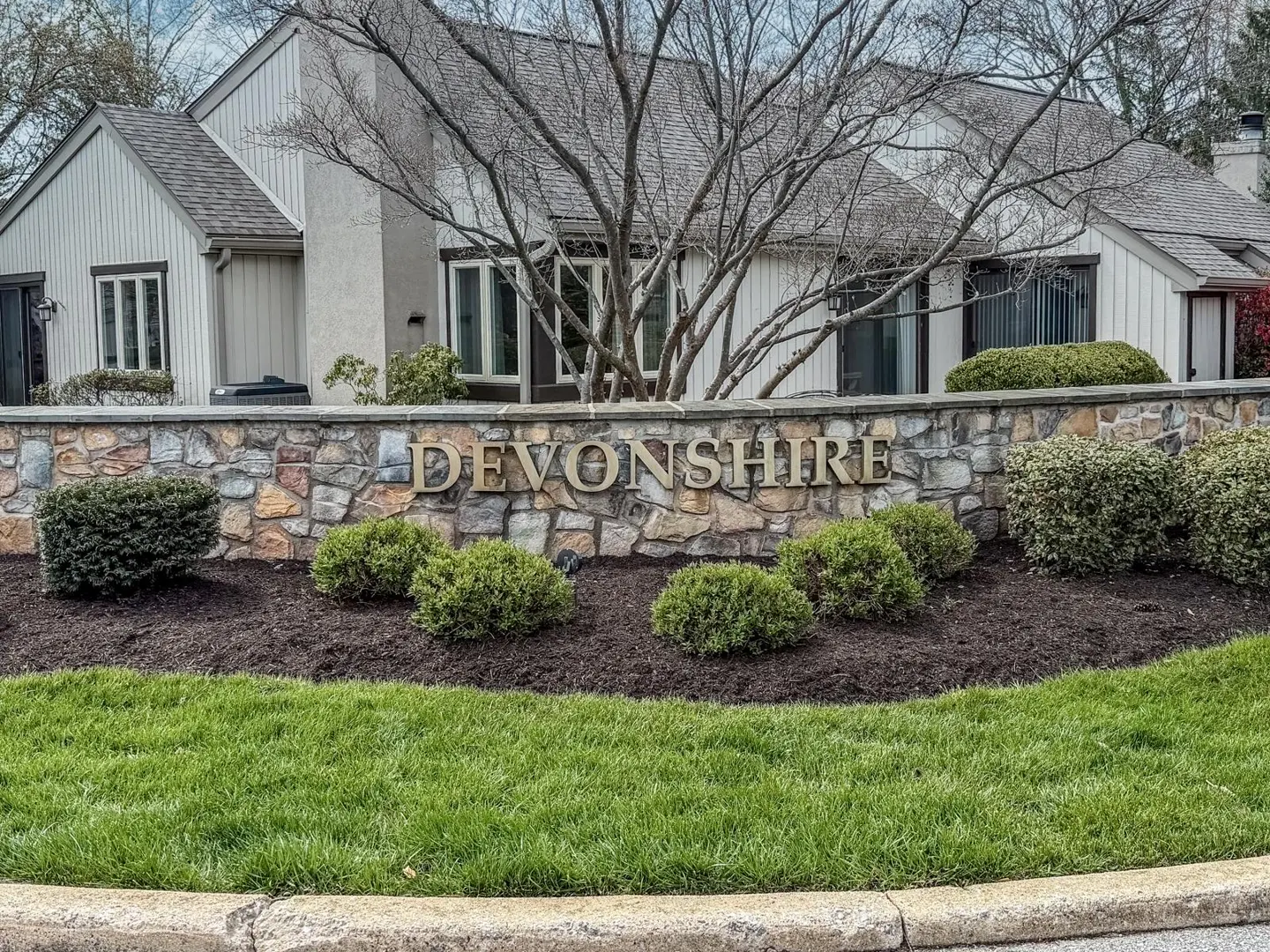 "Devonshire" stone sign with green bushes and grass in front of a beige house with a gray roof and bare trees.