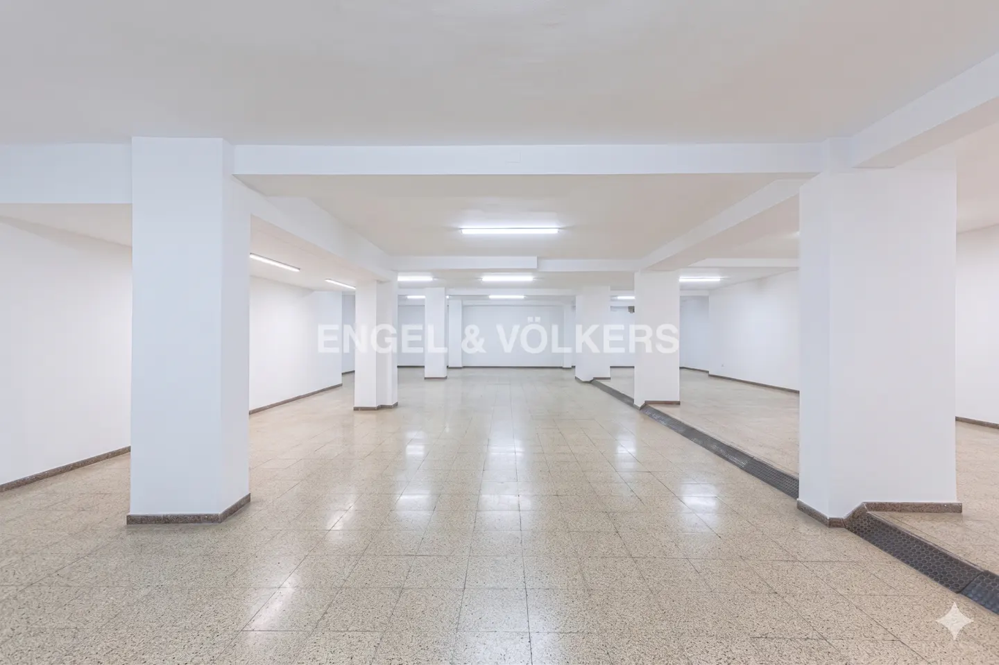 Bright, empty commercial space with white walls, columns, and tile floor. Engel & Völkers logo visible in the background.