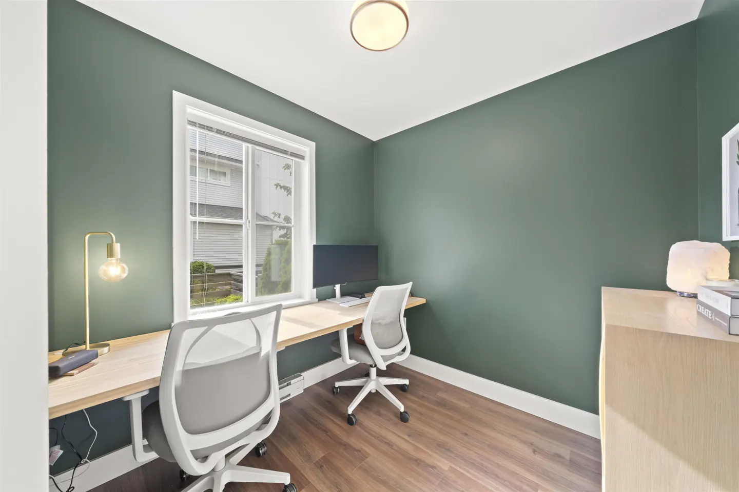 A home office with green walls, wood floors, and a long desk with two chairs. A window provides natural light.