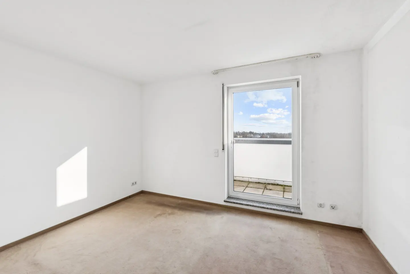 Bright, empty room with white walls and a beige floor. A glass door leads to a balcony with a view of a blue sky.