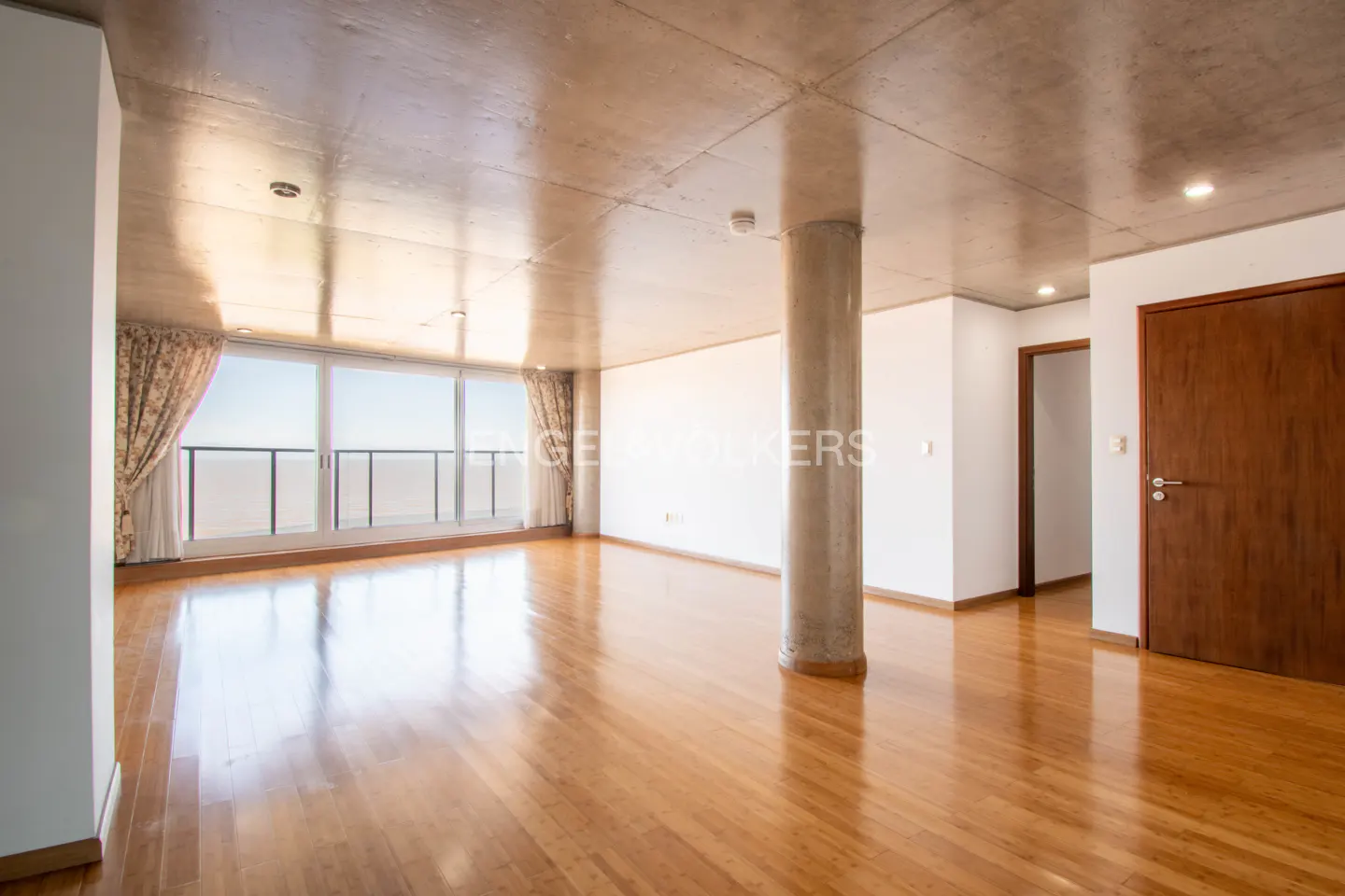 Spacious, bright room with hardwood floors, concrete ceiling, and a large window with ocean view. A concrete pillar stands in the center.