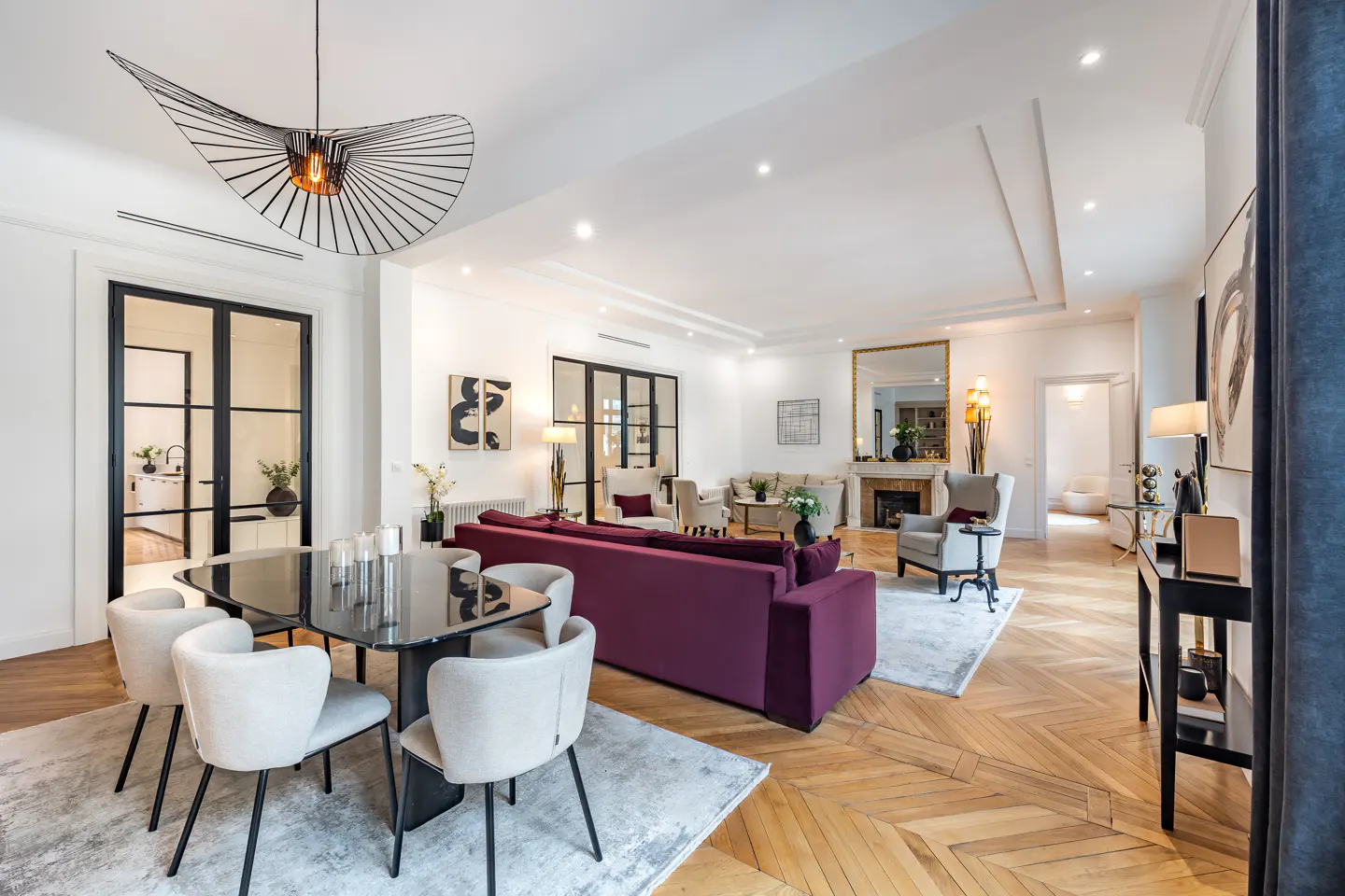 Spacious living and dining room with a deep purple sofa, glass dining table, cream chairs, and herringbone wood floors.