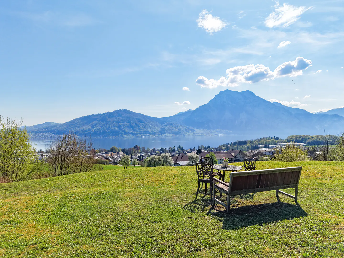 Scenic view of a green lawn with a bench and table set overlooking a lake, town, and mountains under a blue sky.