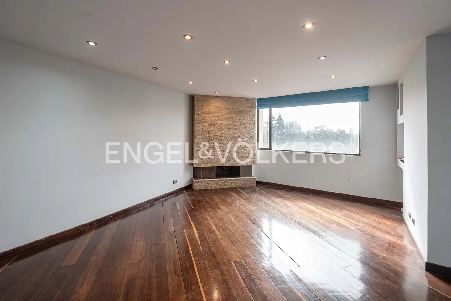 Bright, empty room with hardwood floors, a stone fireplace, and a large window with a blue shade. Spotlights illuminate the ceiling.
