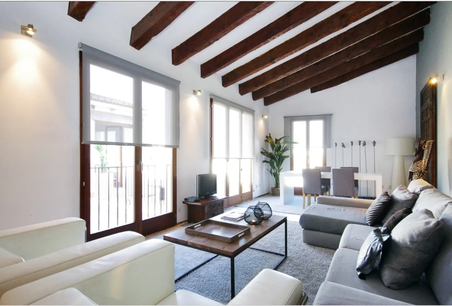 Bright living room with exposed wooden ceiling beams, white walls, gray sofa, coffee table, and large windows with blinds.