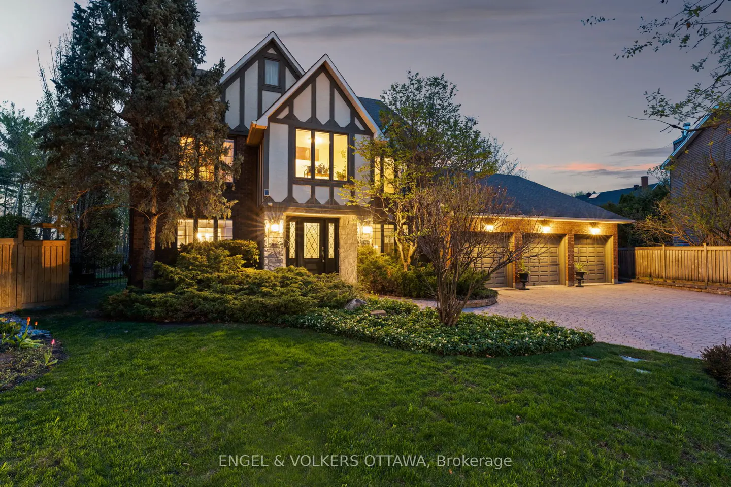 A two-story Tudor-style house with a three-car garage at dusk. The house has a green lawn and manicured landscaping. Interior lights are on.