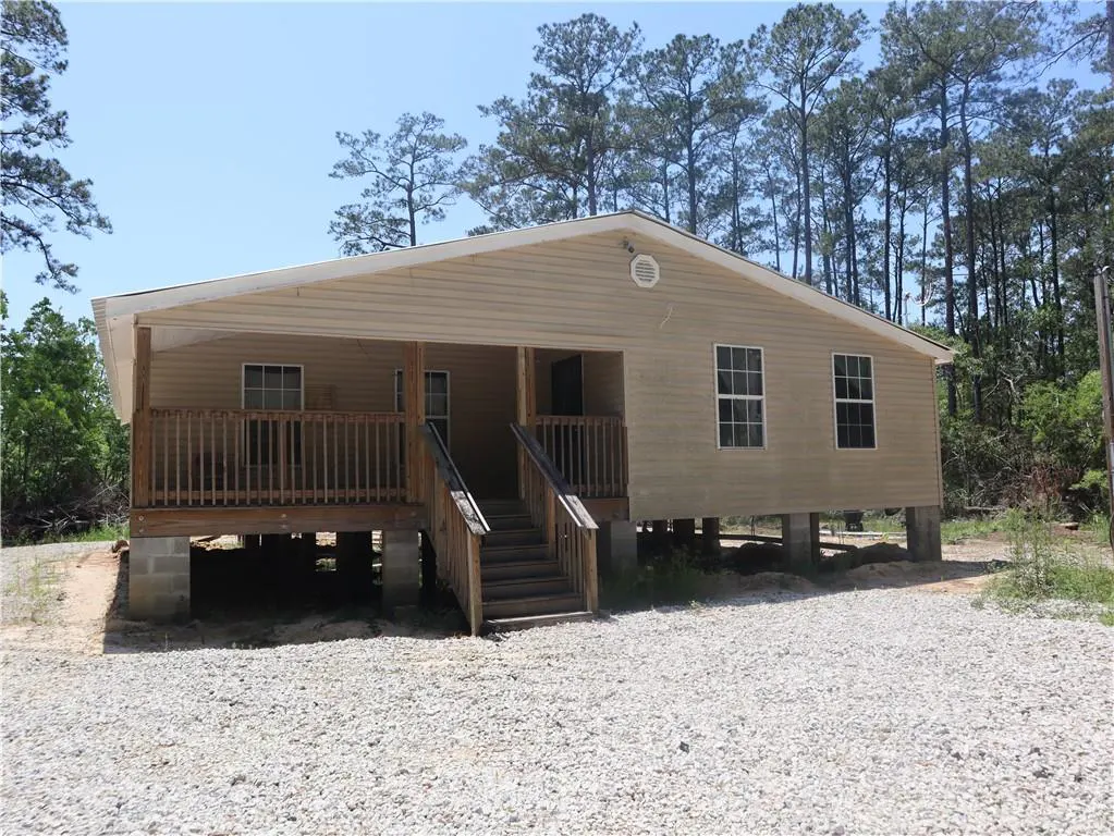 Tan house on concrete blocks with a wooden porch and stairs, surrounded by gravel and trees.