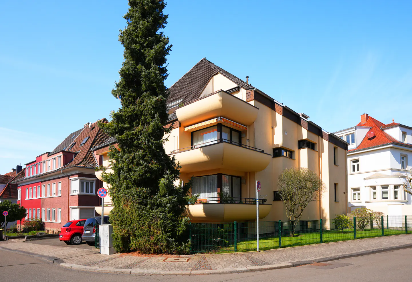 A three-story apartment building with rounded balconies, a tall evergreen tree, and other buildings.