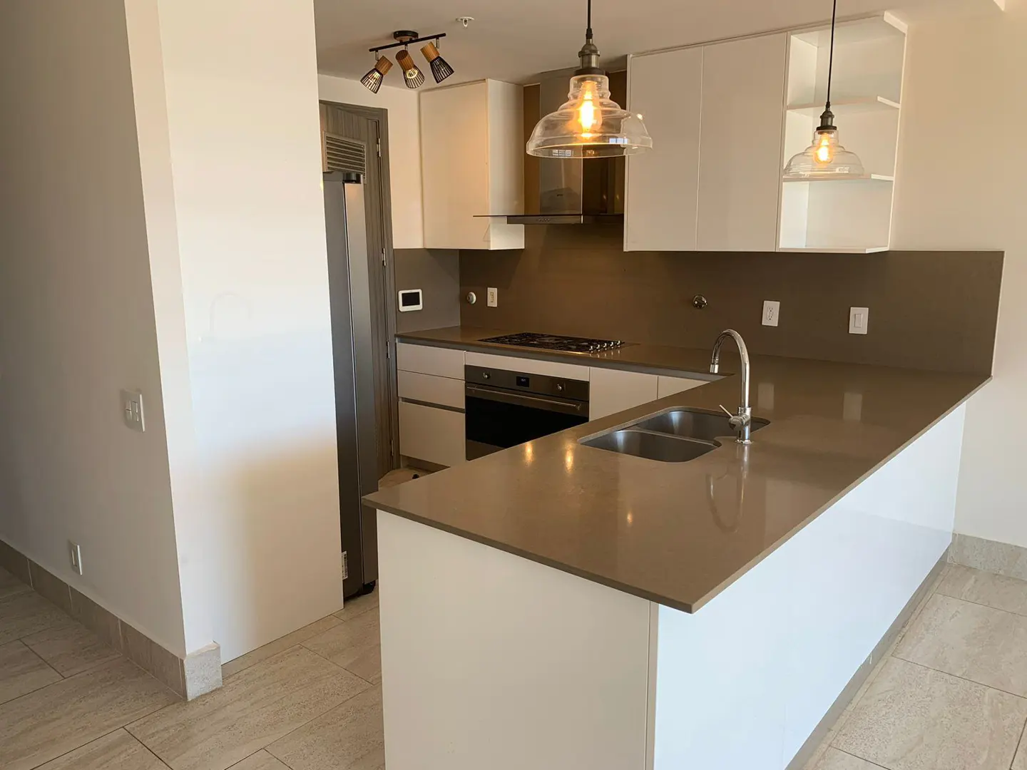 A modern kitchen with white cabinets, stainless steel appliances, and a gray countertop island with a sink. Two pendant lights hang above.