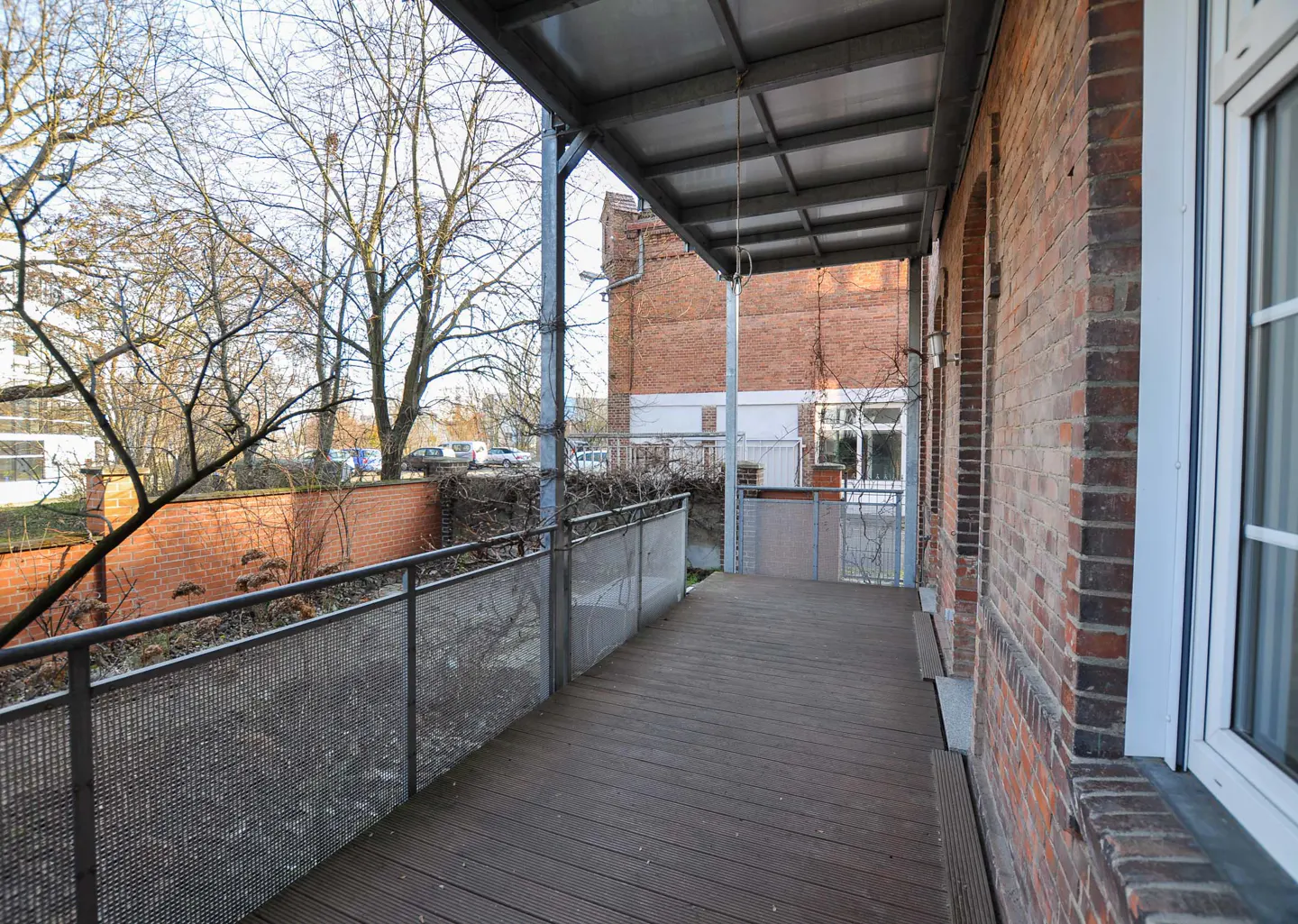 A covered balcony with a wood floor and metal railing overlooks a brick wall and bare trees. A brick building is visible in the background.