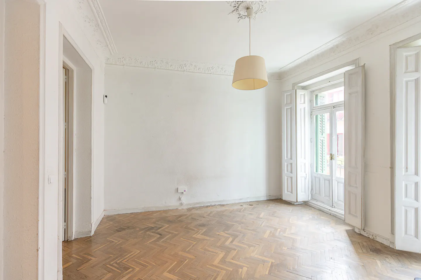 Bright, empty room with herringbone wood floor, white walls, and ornate trim. A tan pendant light hangs from the ceiling. Open white doors lead to a balcony.