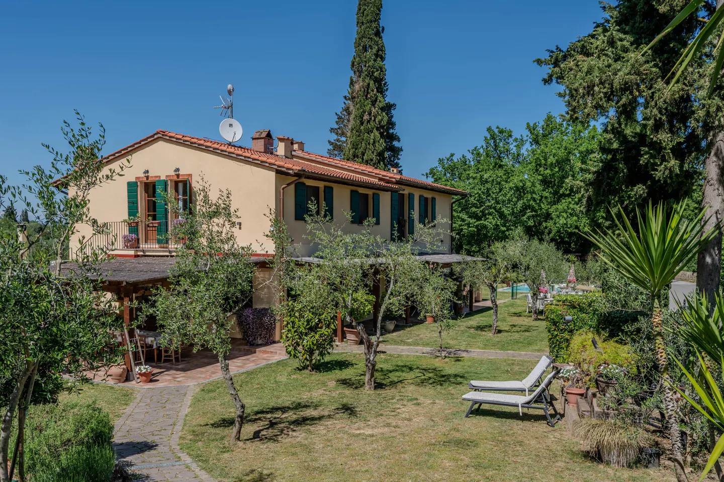 Exterior view of a two-story tan house with green shutters, a red tile roof, and a lush green lawn with trees and lounge chairs.