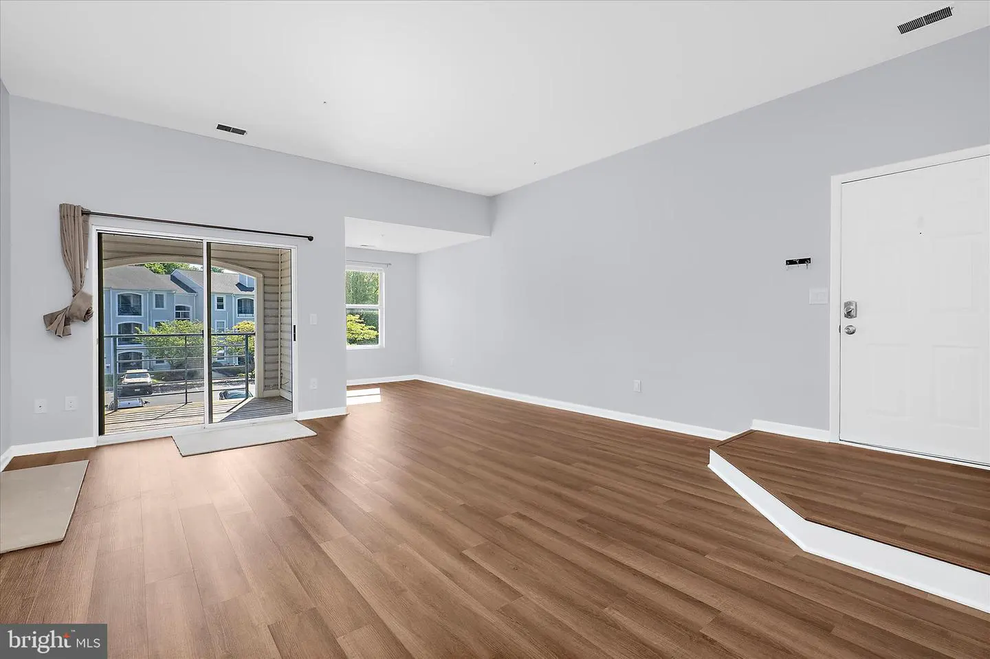 A bright, empty living room with wood floors, light blue walls, and a sliding glass door to a balcony.