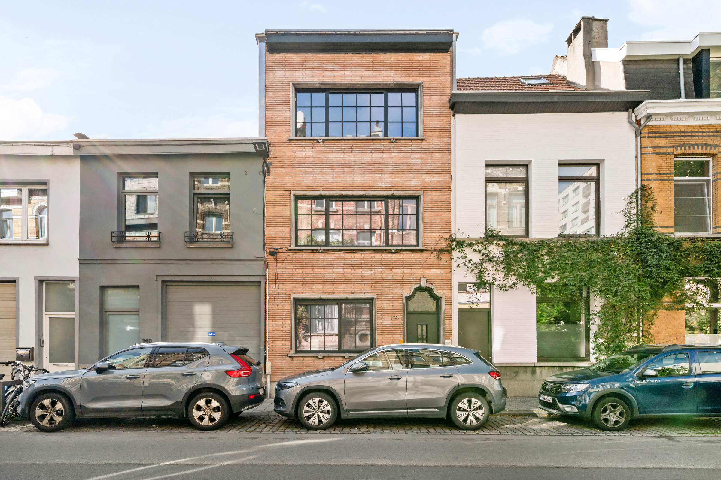 A narrow brick townhouse sits between a grey building and a white building with ivy, with cars parked on the street. [AI-generated text]
