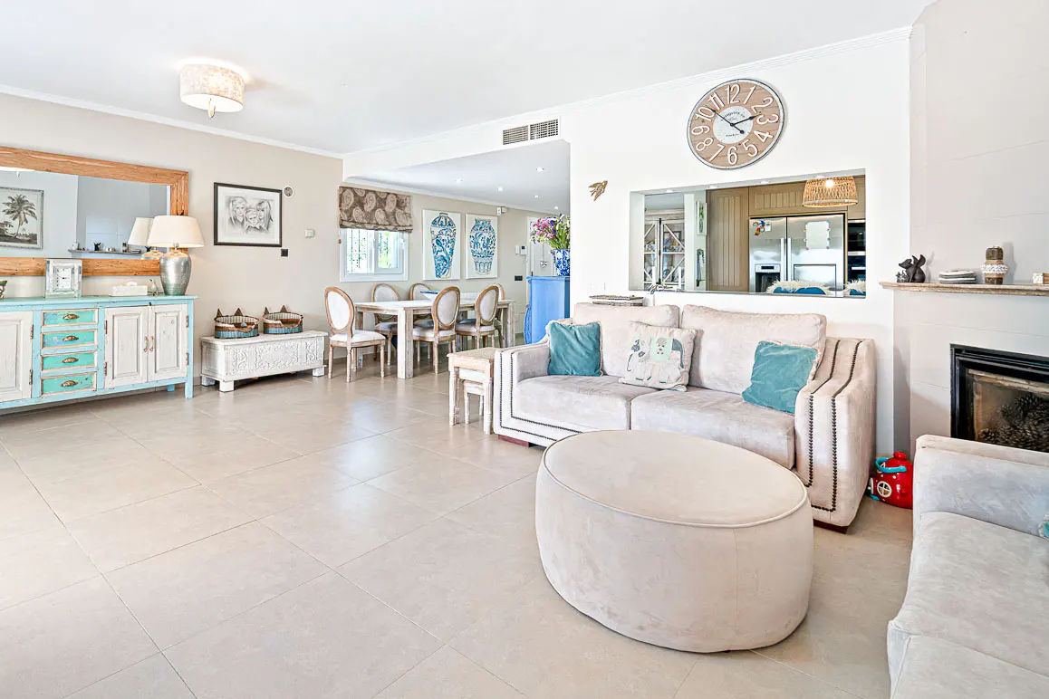 A bright, open-concept living room with a beige sofa, round ottoman, and a dining area in the background. A large clock hangs on the wall.