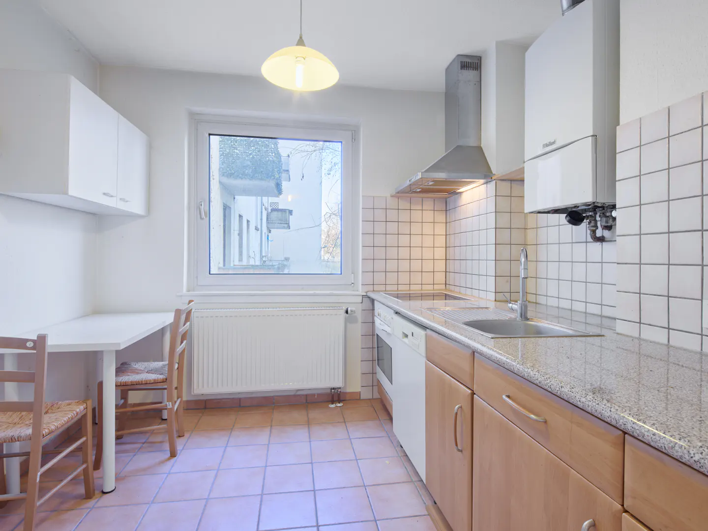A bright kitchen with light wood cabinets, white appliances, and a table with two chairs. A window overlooks a building.