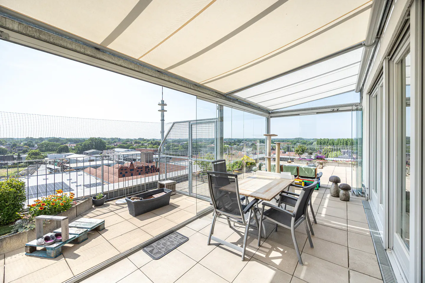 Covered balcony with a dining set, potted plants, and a glass enclosure overlooking a town with a distant radio tower.