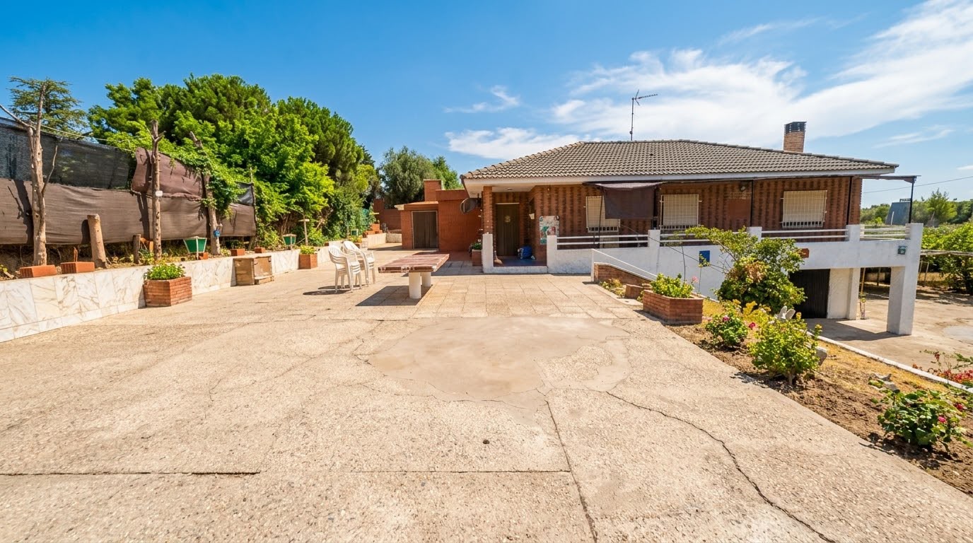 A red brick house with a tiled roof features a large, cracked concrete patio, white chairs, and a table under a blue sky. [AI-generated text]