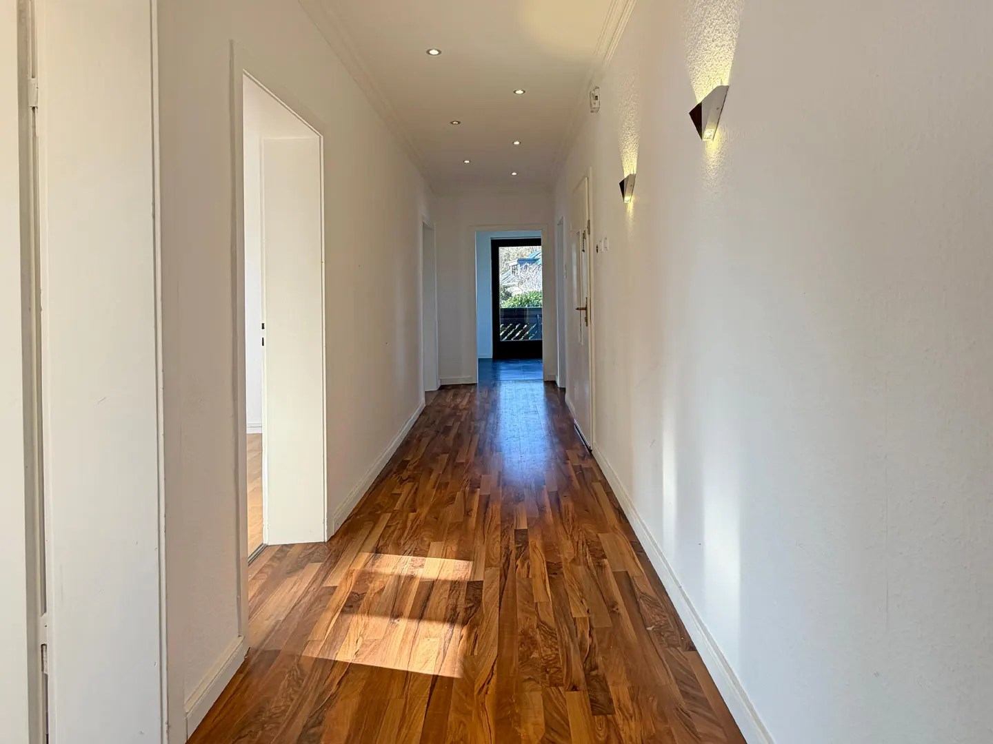A bright hallway with white walls, wood floors, and recessed lighting. Doors line the hall, leading to rooms and a balcony with a view.