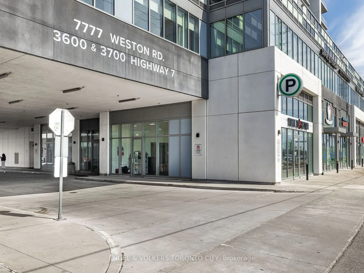 Exterior view of a modern building with retail spaces and parking entrance. Signage indicates addresses and parking.