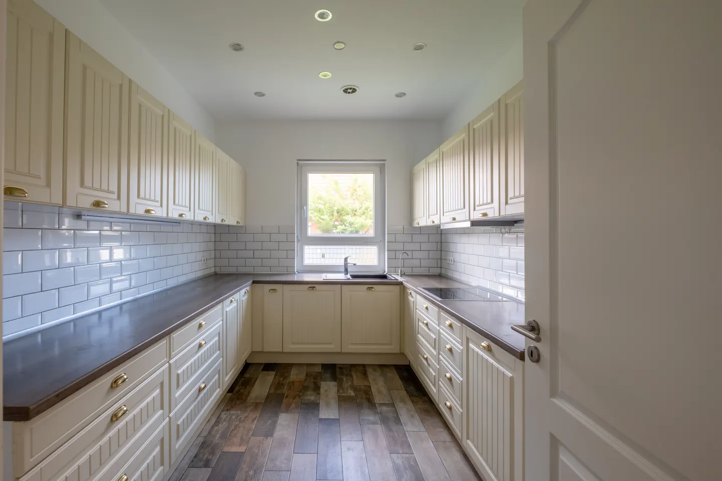 A bright kitchen with cream cabinets, dark countertops, and white subway tile. A window is centered above the sink.