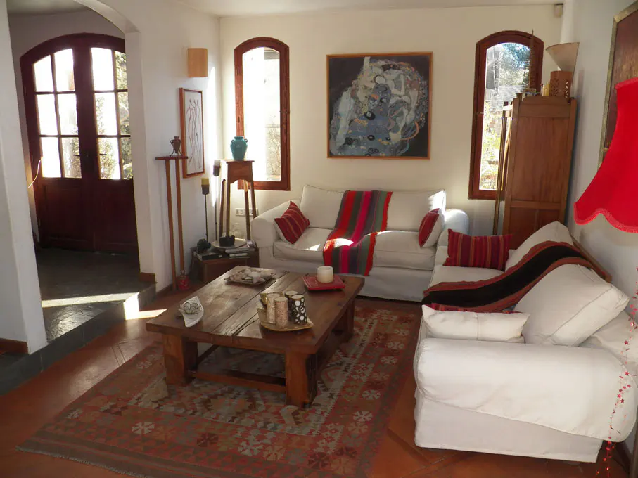 Living room with white sofas, wood table, and patterned rug. Artwork hangs above the sofa, and light streams through arched windows.