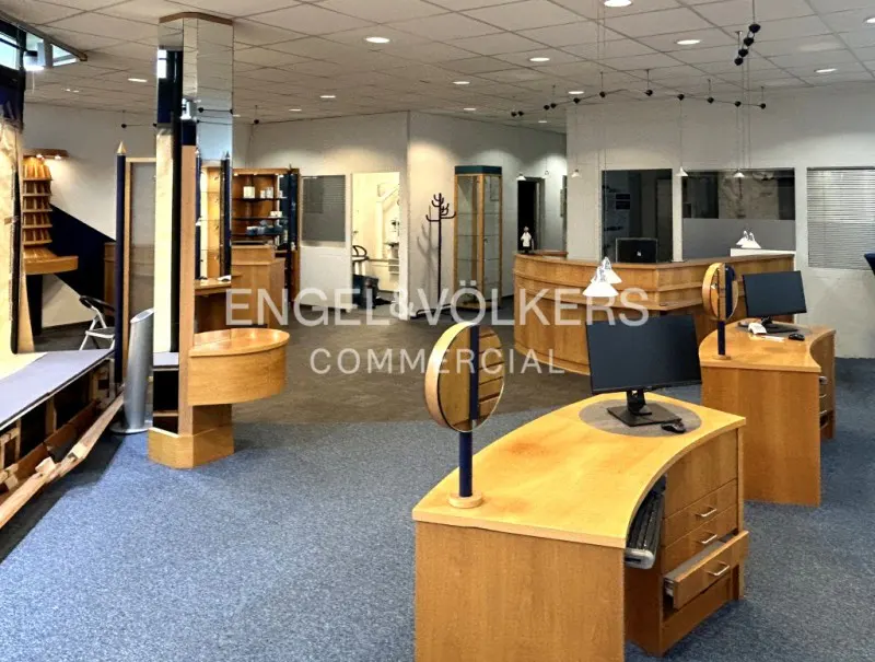 A commercial space with light wood desks, blue carpet, and white walls. The Engel & Völkers logo is visible in the center.