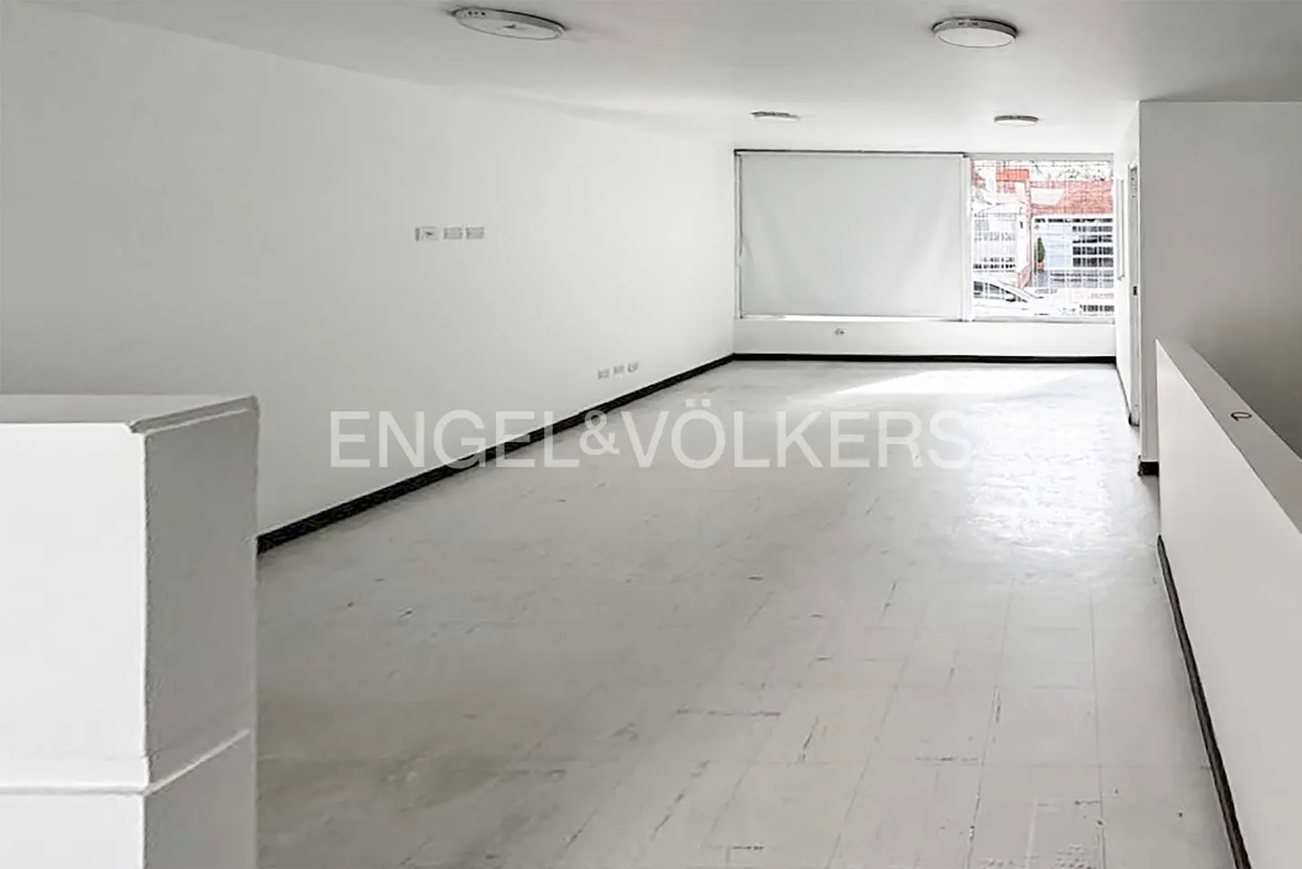 Empty white room with a large window and white roller blind. The Engel & Völkers logo is in the center of the image.