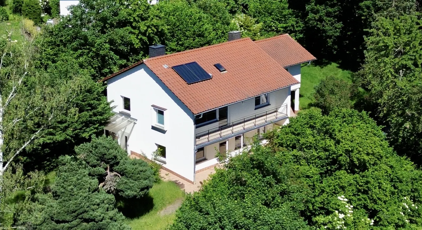 Two-story white house with a red tile roof and solar panels, surrounded by green trees.