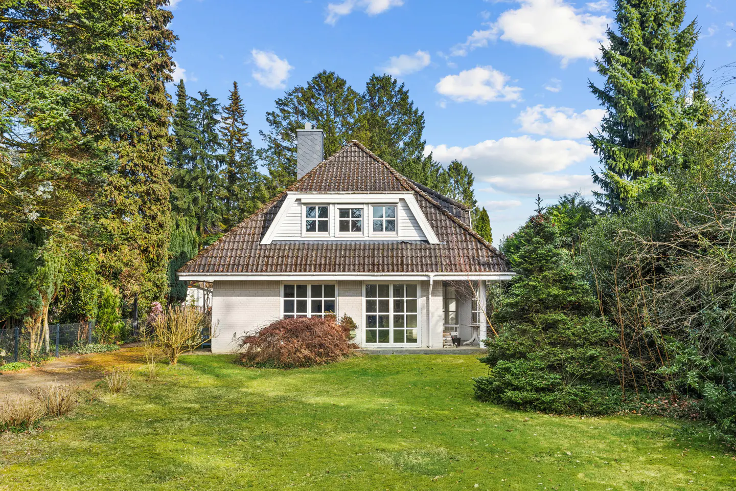 A white house with a brown tile roof sits on a green lawn surrounded by tall trees.