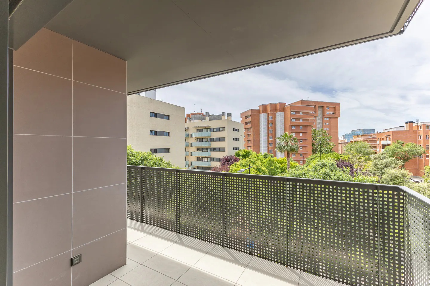 View from a balcony with a dark metal railing overlooking city buildings and green trees.