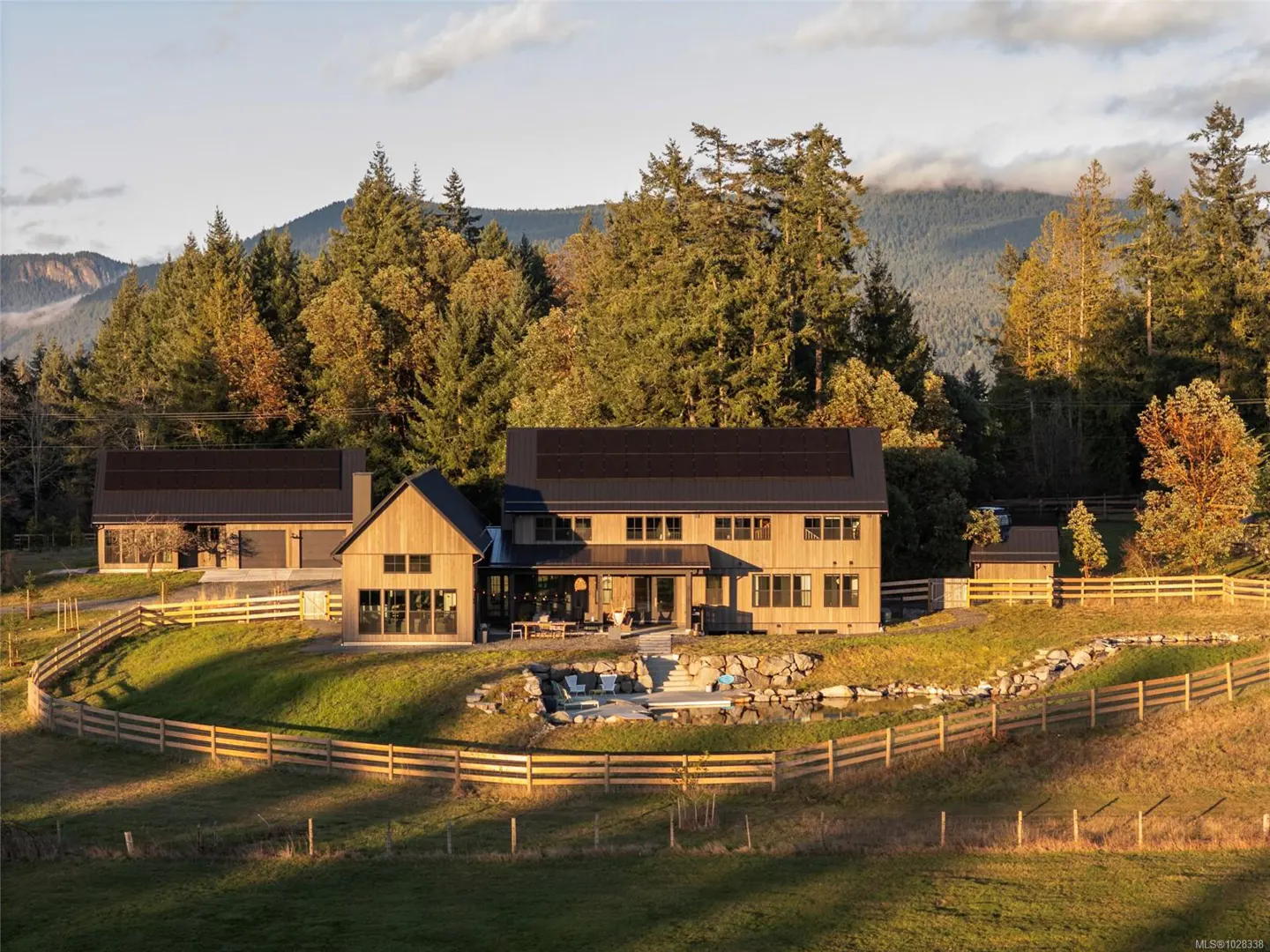A modern farmhouse with a dark roof sits on a grassy hill surrounded by a wooden fence and trees.