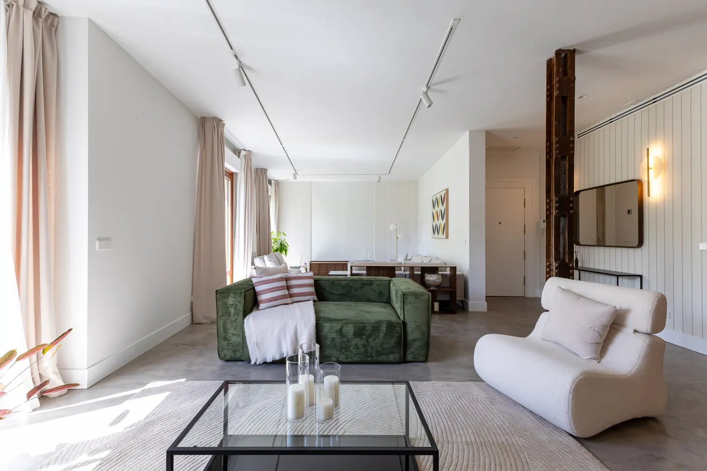 Bright living room with a green sofa, white chair, and glass coffee table on a light rug. Track lighting illuminates the space.