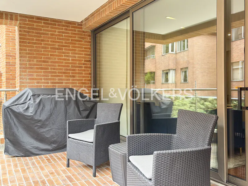 Balcony with brick walls, two wicker chairs with white cushions, and a covered grill. Sliding glass doors open to a view of another brick building.