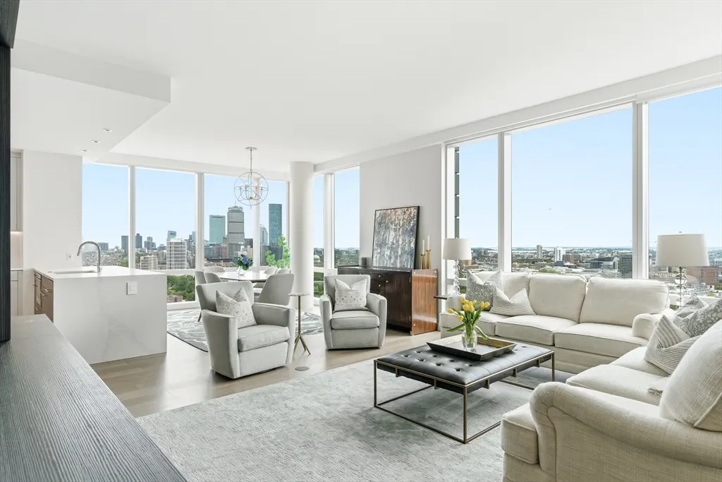 Bright, modern living room with floor-to-ceiling windows overlooking a city skyline. Gray armchairs and a white sofa surround a black leather ottoman.