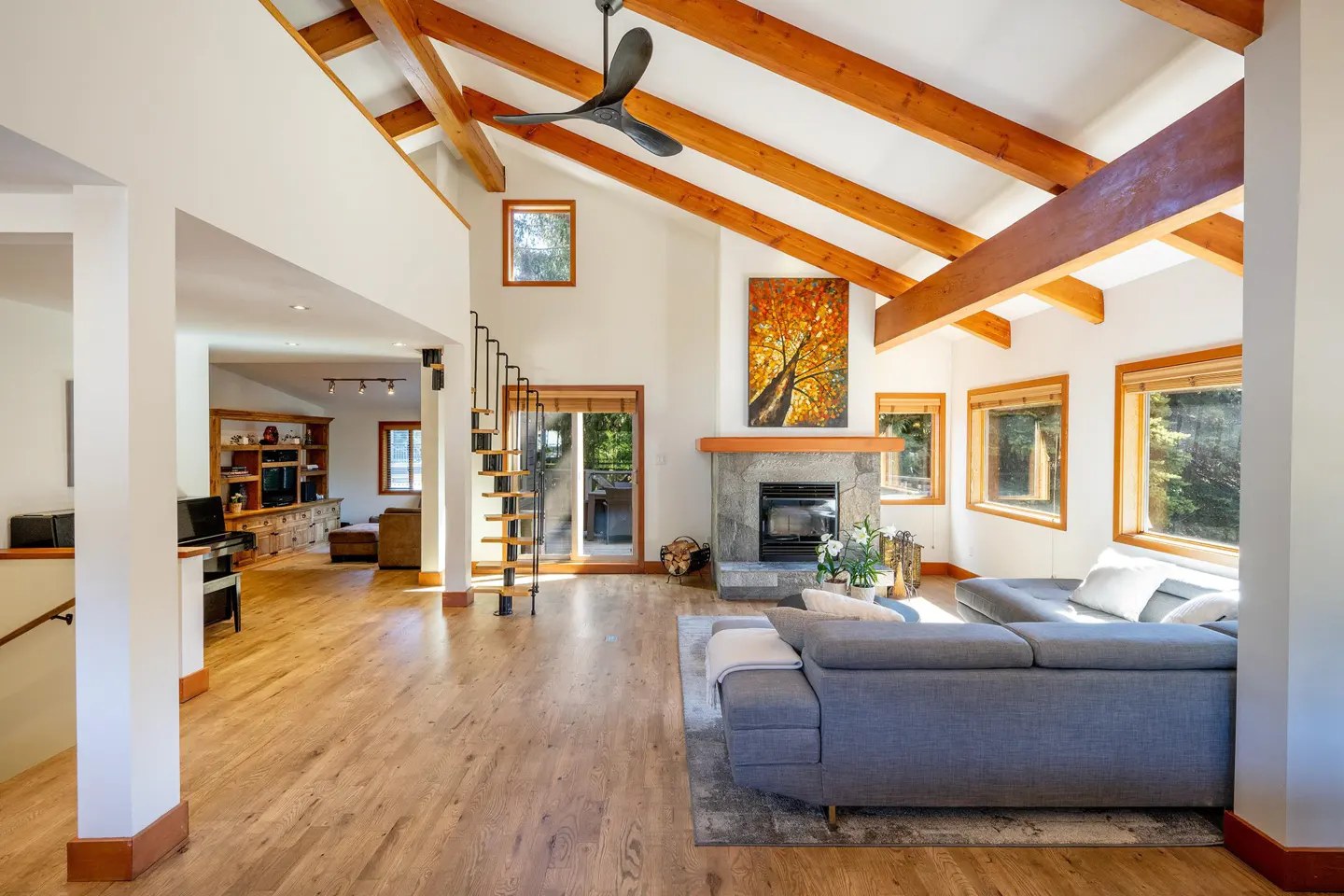 Bright living room with wood floors, exposed beams, and a gray sectional sofa. A stone fireplace and spiral staircase add character.