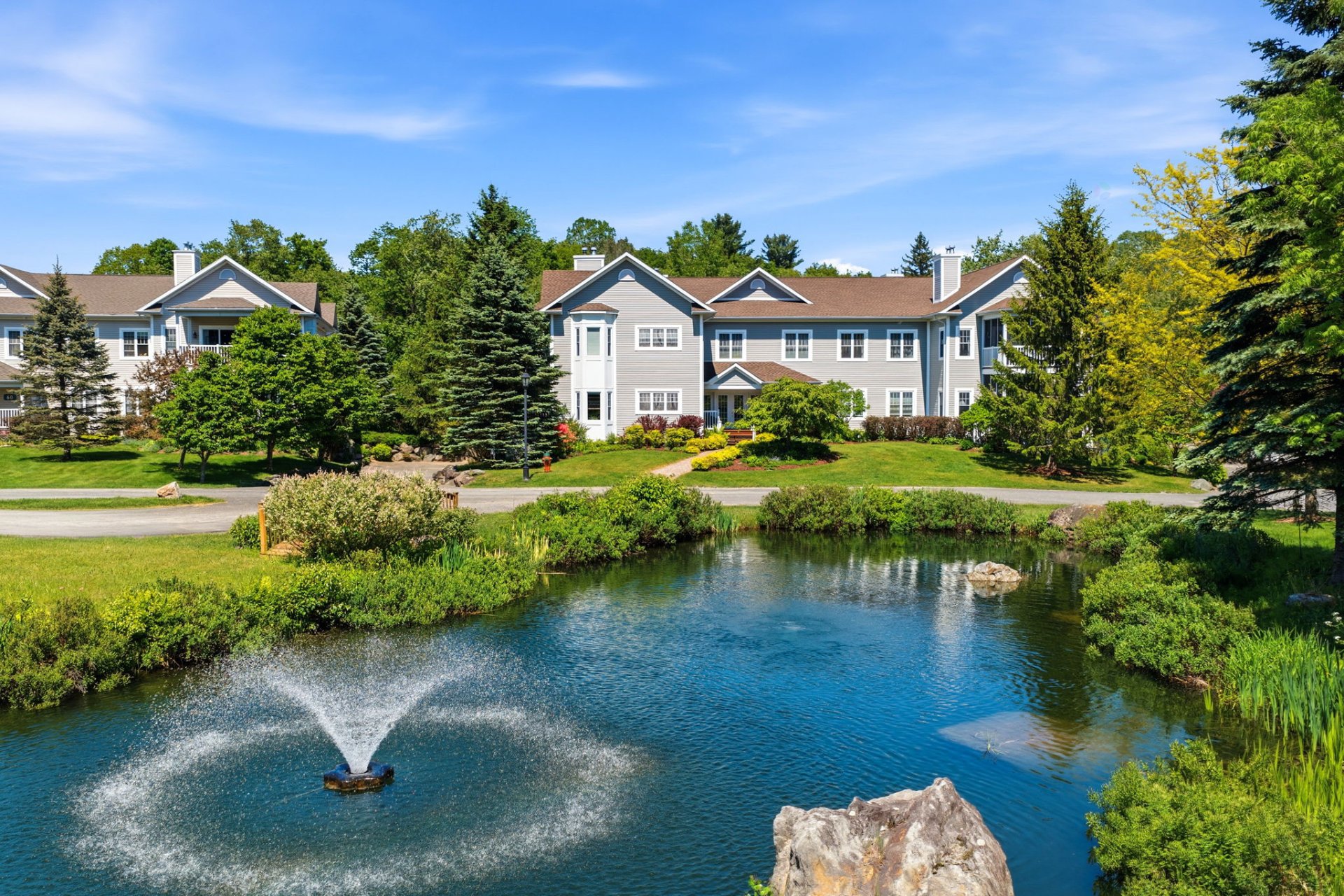 A pond with a fountain in the foreground, set before a large, light-gray multi-unit residential building on a sunny day.