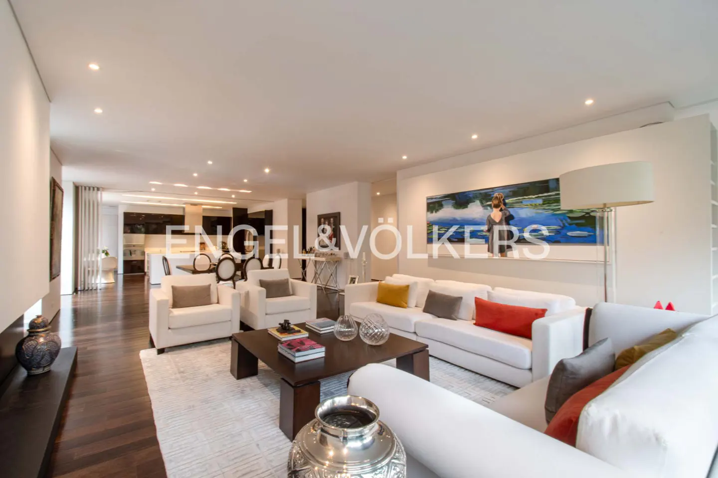 Bright, open-plan living room with white sofas, armchairs, and a dark wood floor. A large painting hangs on the wall above the sofa.