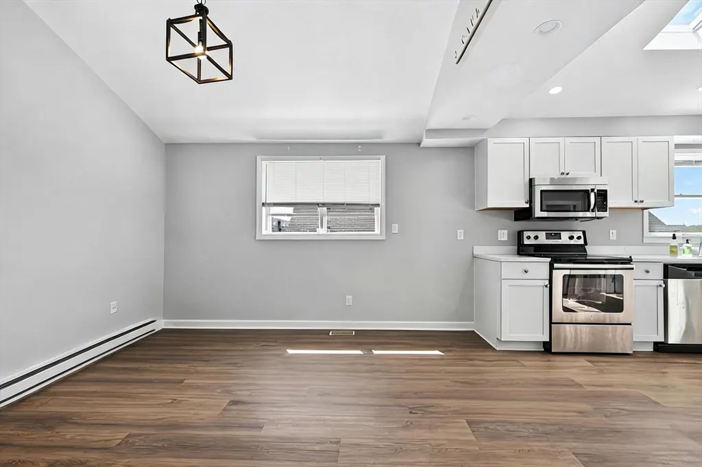 A bright, open-concept kitchen and dining area with wood floors, gray walls, and white cabinets. Stainless steel appliances are visible.