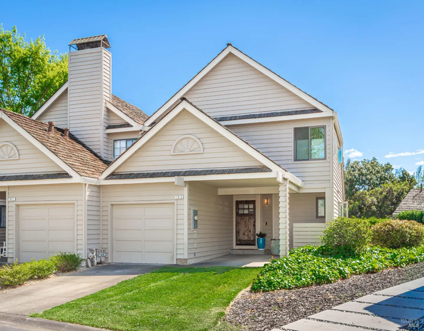 Beige two-story townhouse with a brown roof, a garage, and a well-manicured lawn on a sunny day.