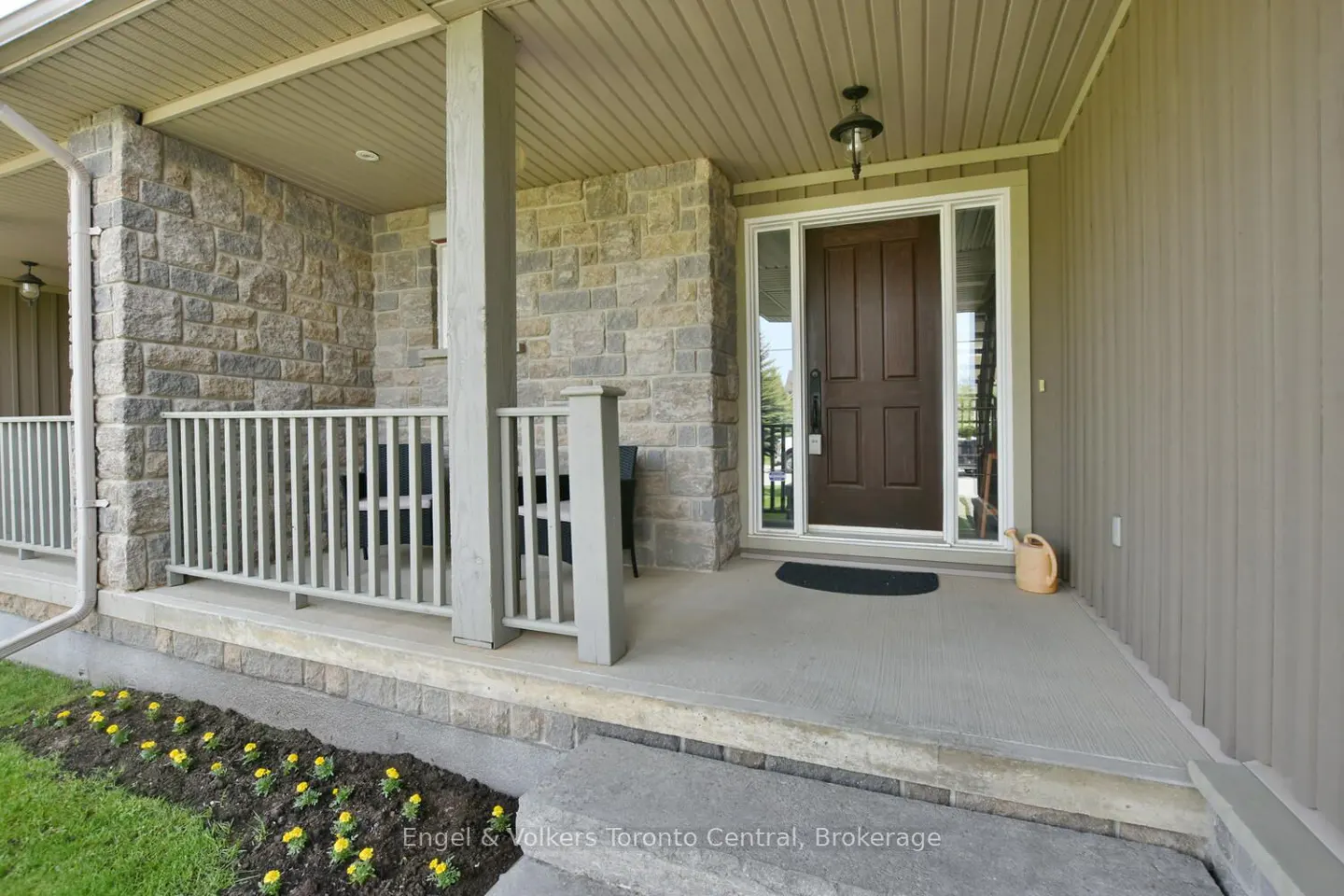 Covered front porch with stone facade, dark brown door, light grey railing, and a small flower bed with yellow flowers.