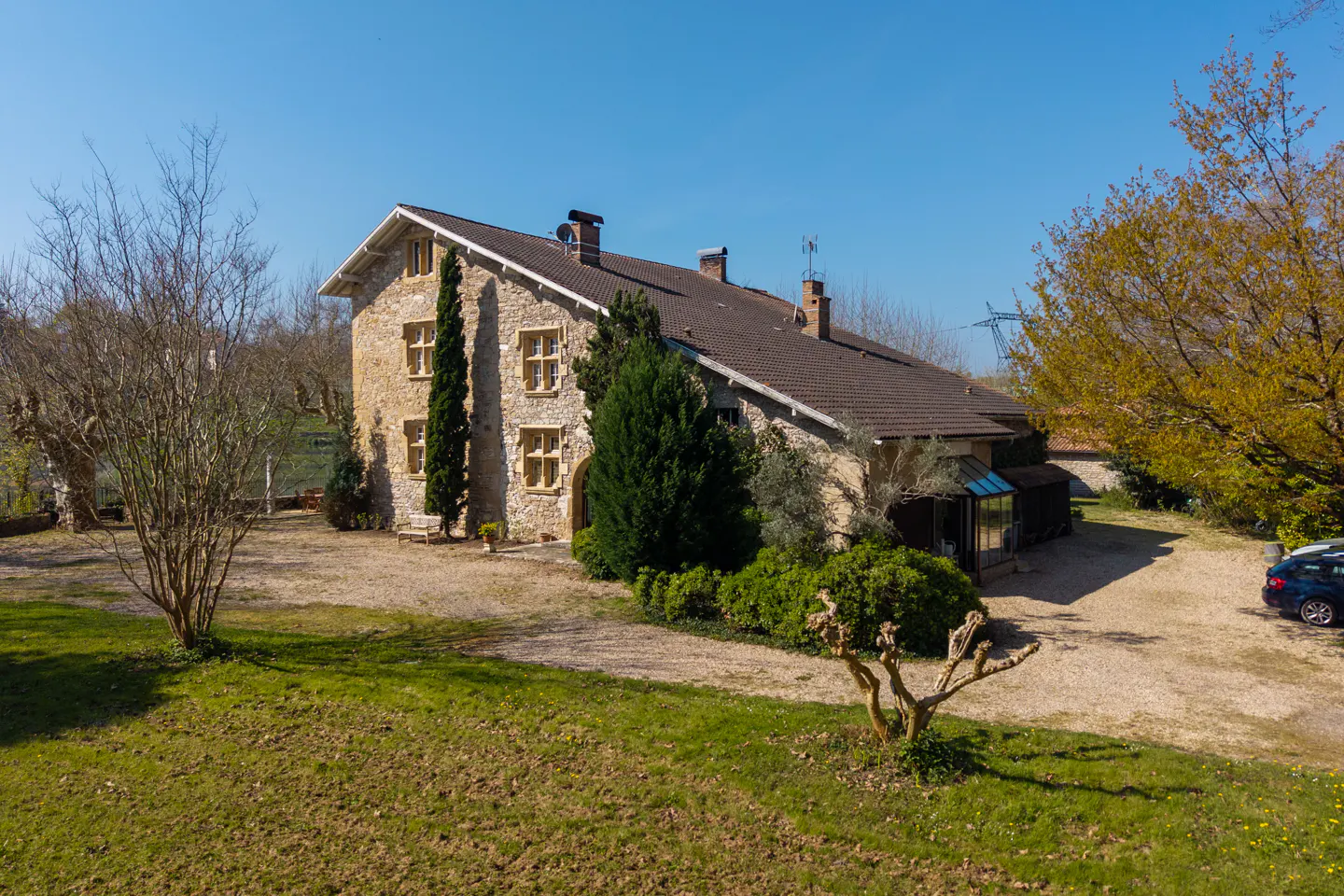 Stone house with brown roof, green lawn, and gravel driveway on a sunny day.