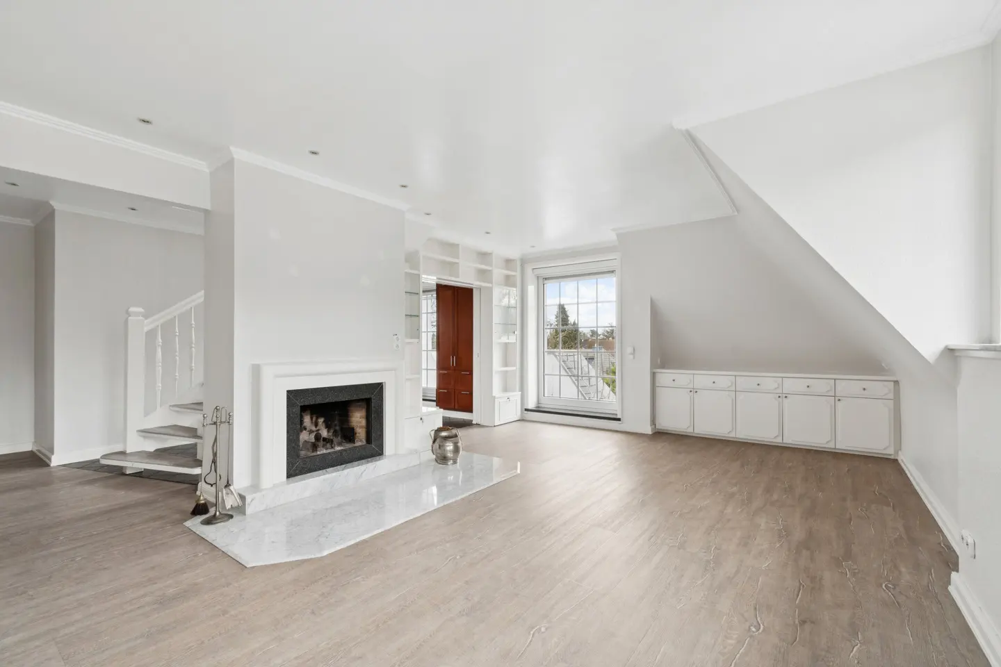 Bright, airy living room with a white fireplace, marble hearth, and wood floors. A staircase and built-in cabinets add architectural detail.
