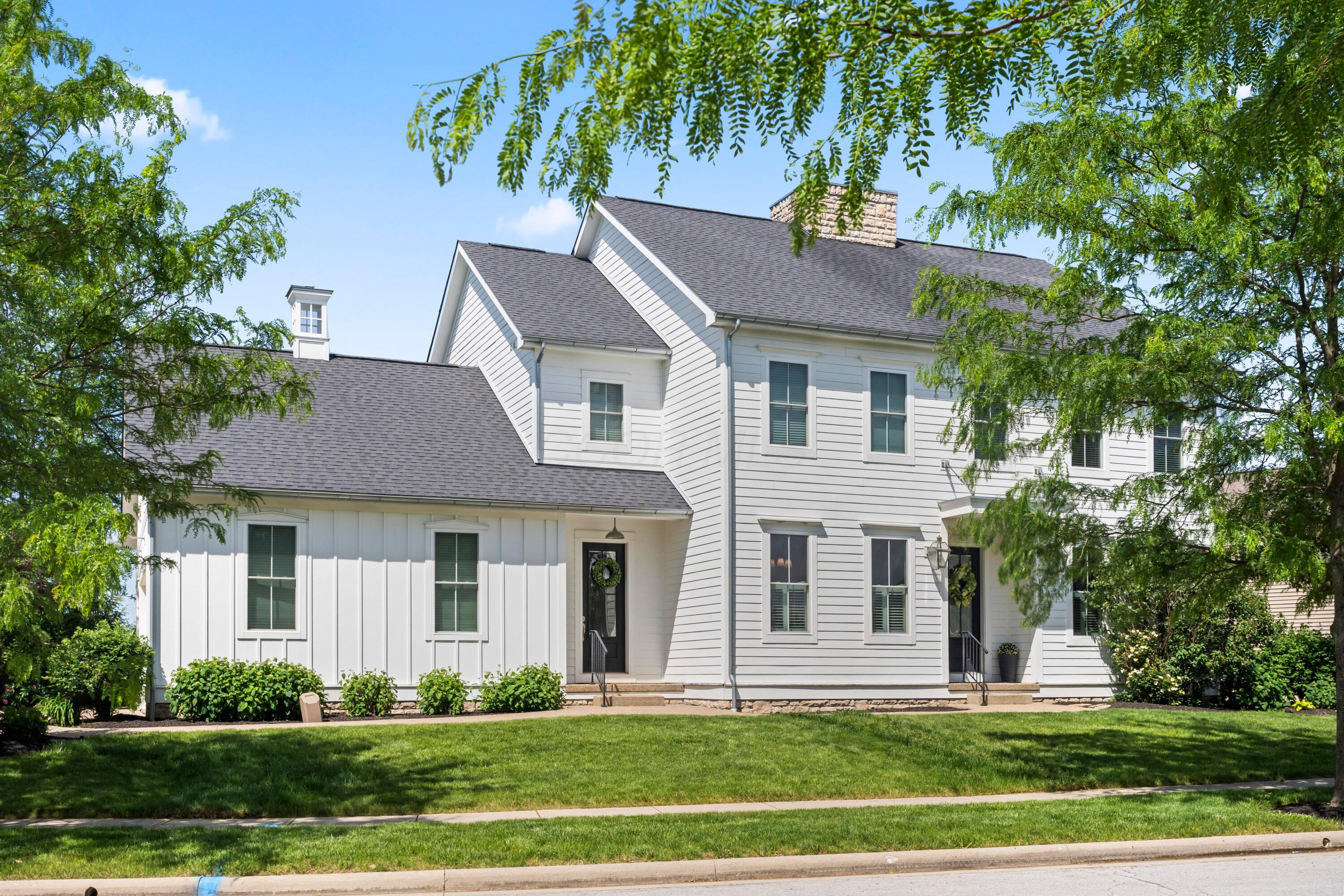 Two-story white farmhouse with a gray roof, chimney, and black doors. Green lawn and trees frame the house against a blue sky.