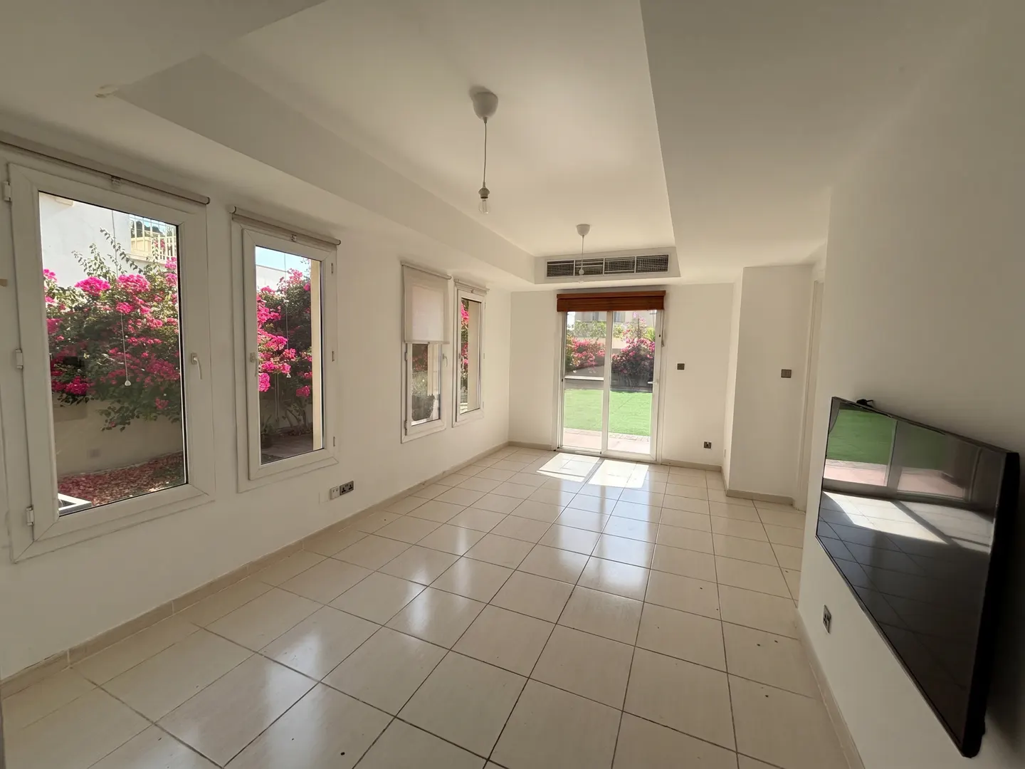 Bright, empty room with white walls, tile floor, and large windows. Pink flowers are visible outside. A TV is mounted on the right wall.