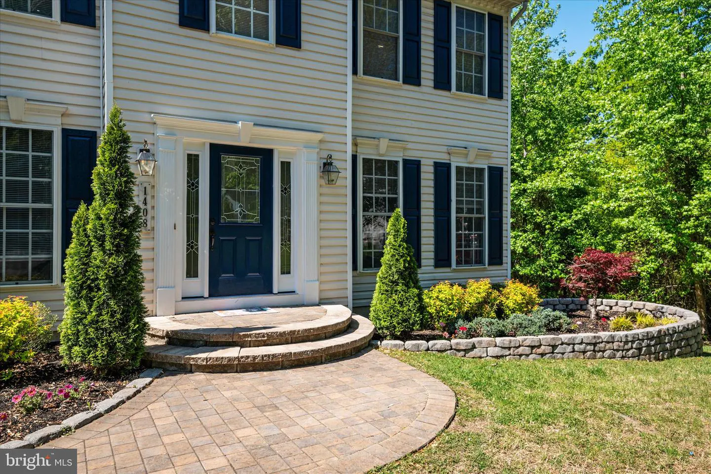 Two-story house with beige siding, blue shutters, and a blue front door with decorative glass. A brick walkway leads to the entrance.