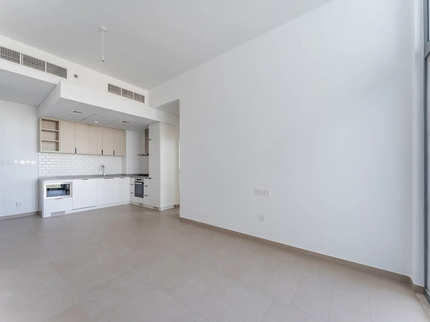 Bright, empty room with white walls and beige tile floor. Kitchen with white cabinets and stainless steel appliances in the background.