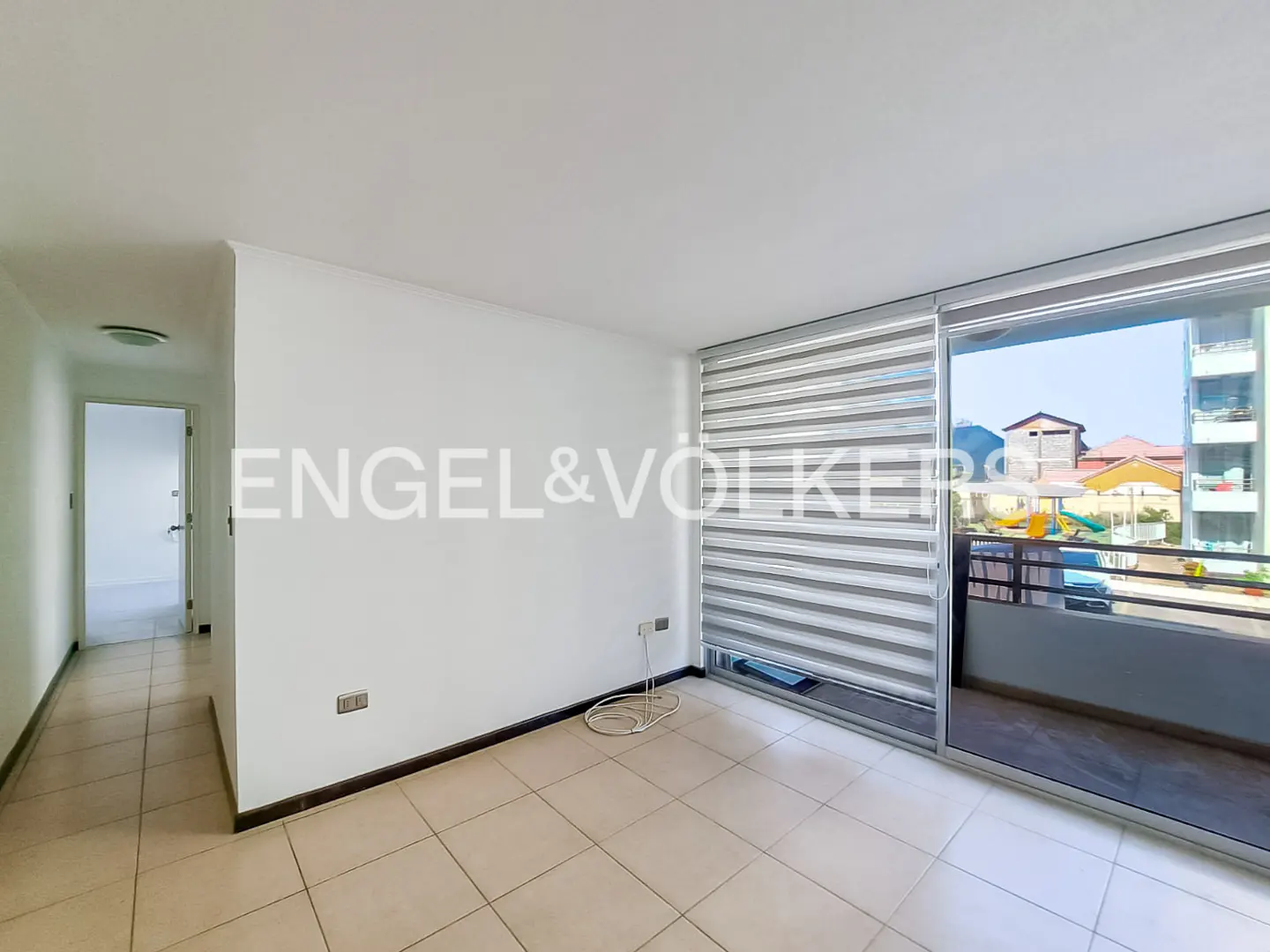 Bright, empty room with beige tile floor, white walls, and a sliding glass door to a balcony. Engel & Völkers logo on the wall.