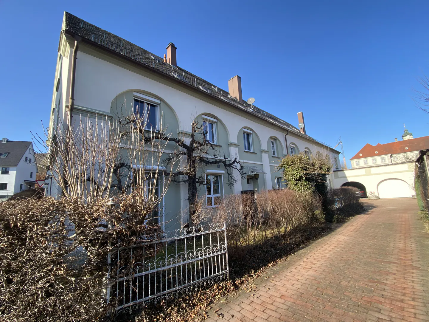 A long, light-colored residential building with arched window frames, a brick driveway, and a metal fence under a blue sky.